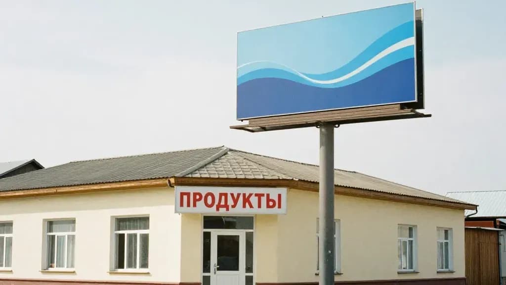 Облыстық қаланың күндізгі көшесі: кіреберісі үстінде маңдайшасы бар дүкен, бұрышта тірекке орнатылған жарнама қалқаны; адам жоқ